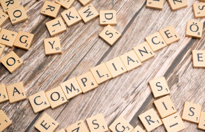 Wooden letter tiles forming the word 'COMPLIANCE' on a rustic wooden background.
