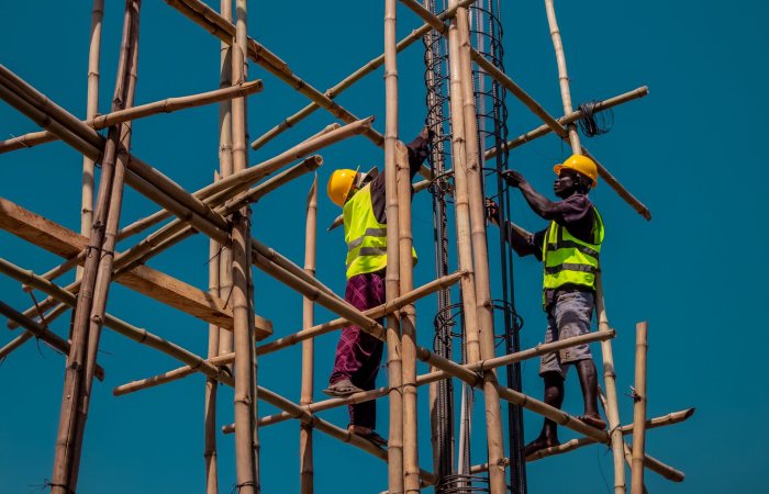 Two construction workers on bamboo scaffolding, wearing safety gear under a clear blue sky.